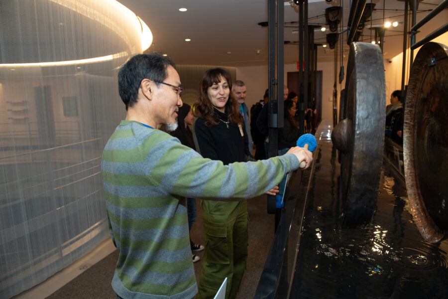 two adults look at a gong hanging above a tub of water, with one person holding a mallet