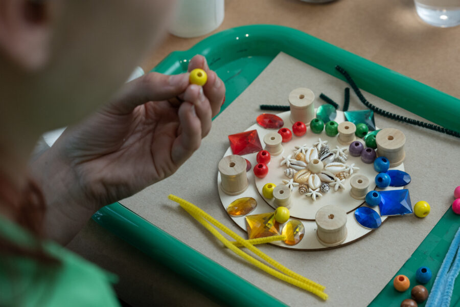 A close up of someone's hand holding a yellow bead. A green tray on a table holds a circle mandala that is made up of colorful beads, wooden beads, shells, and pipe cleaners.