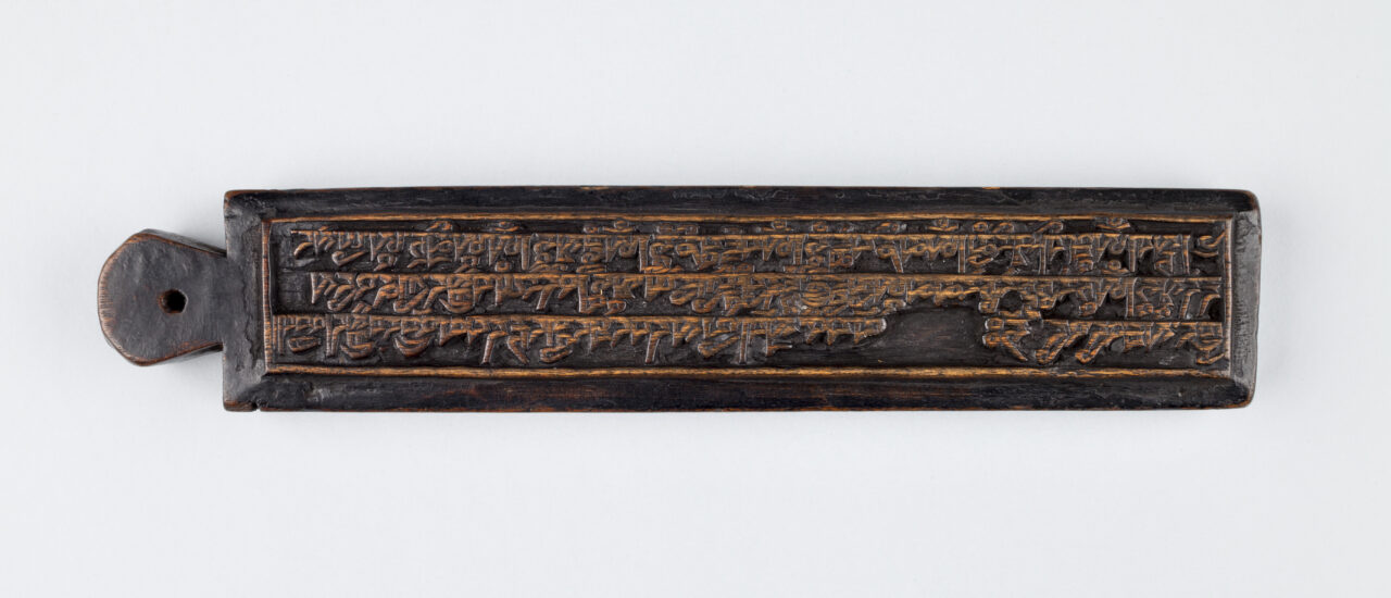 long, rectangular, ink-stained woodblock incised with dense text; handle with hole in center at left