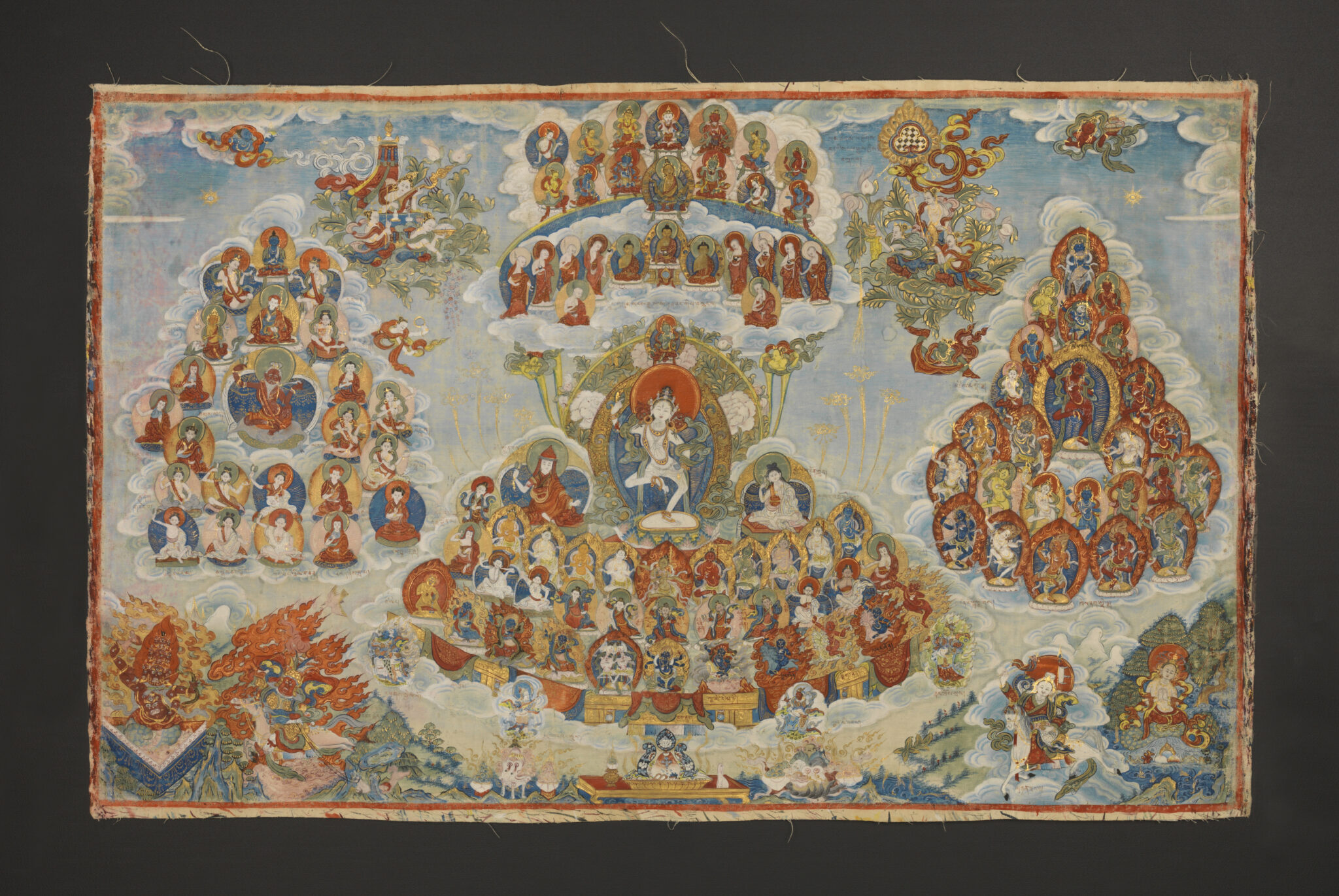 Three clusters of portraits centered on white-skinned deity in dancing pose hover above mountainous landscape