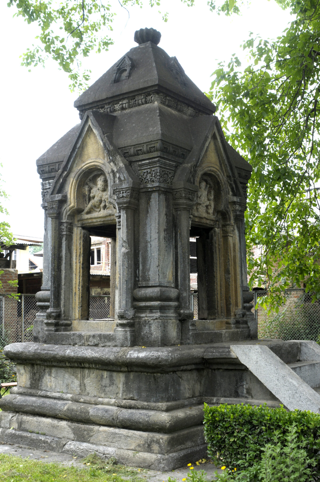 Exterior view of canopied monument in weathered gray and brown stone featuring pointed arches and tiered pyramid roof