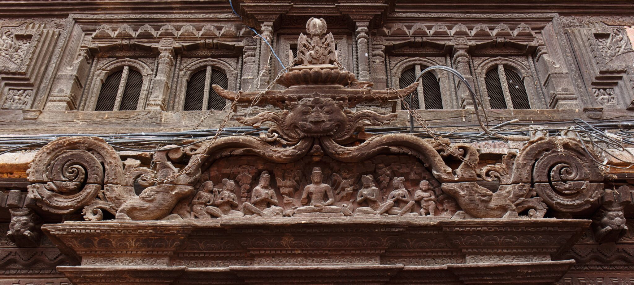 Medium view of densely carved brown wooden lintel beneath row of small windows; features scene with seven seated figures framed by scrollwork