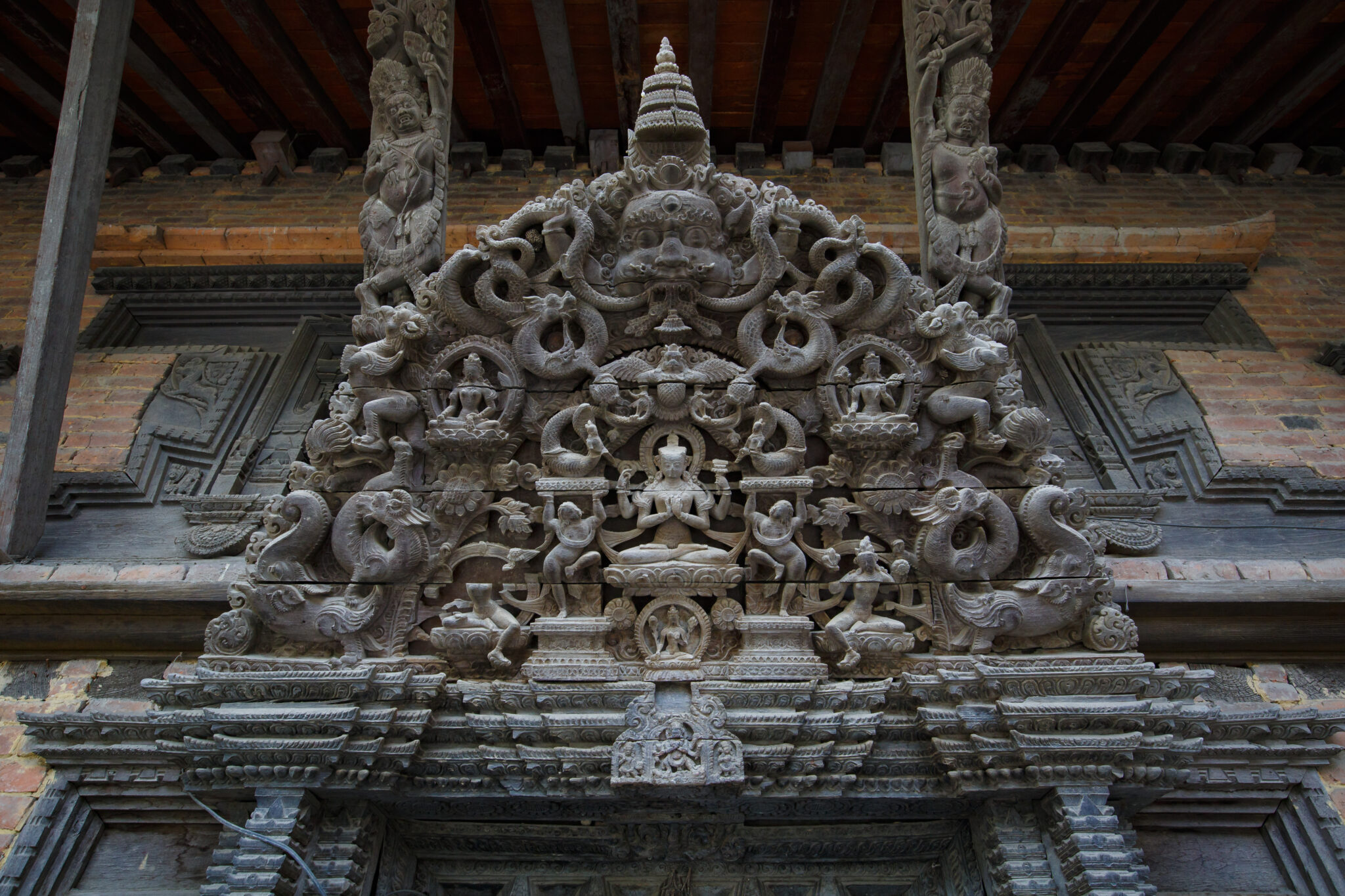 Lavishly carved semicircular gray-brown woodwork with seated Buddha at center; mounted on densely patterned lintel