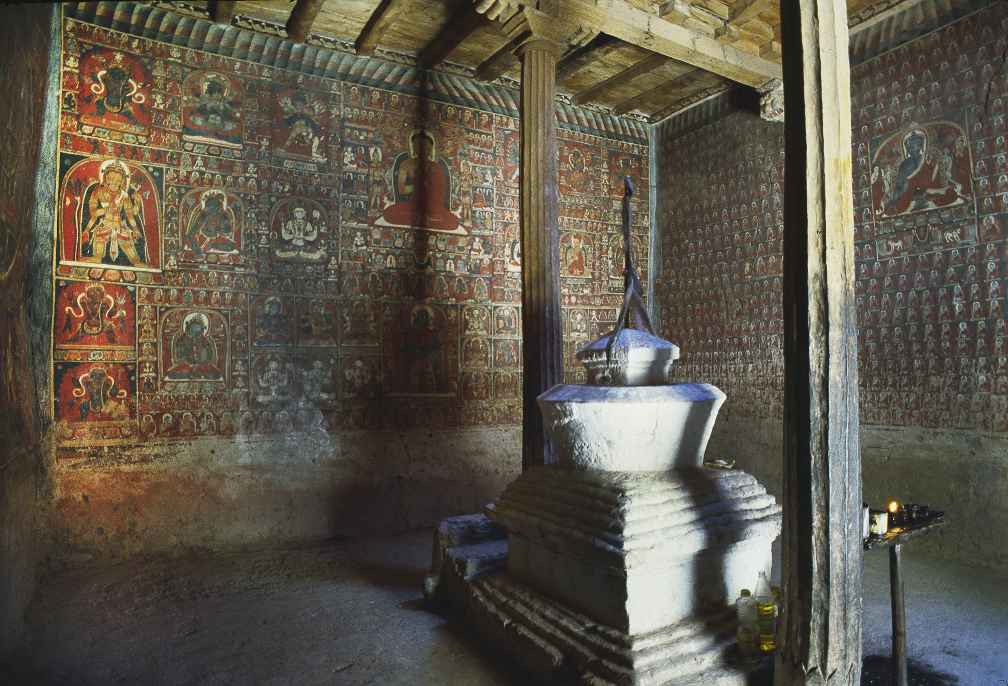 Mural depicting dozens of figures arranged in irregular grid pattern illuminated in half-light behind chorten