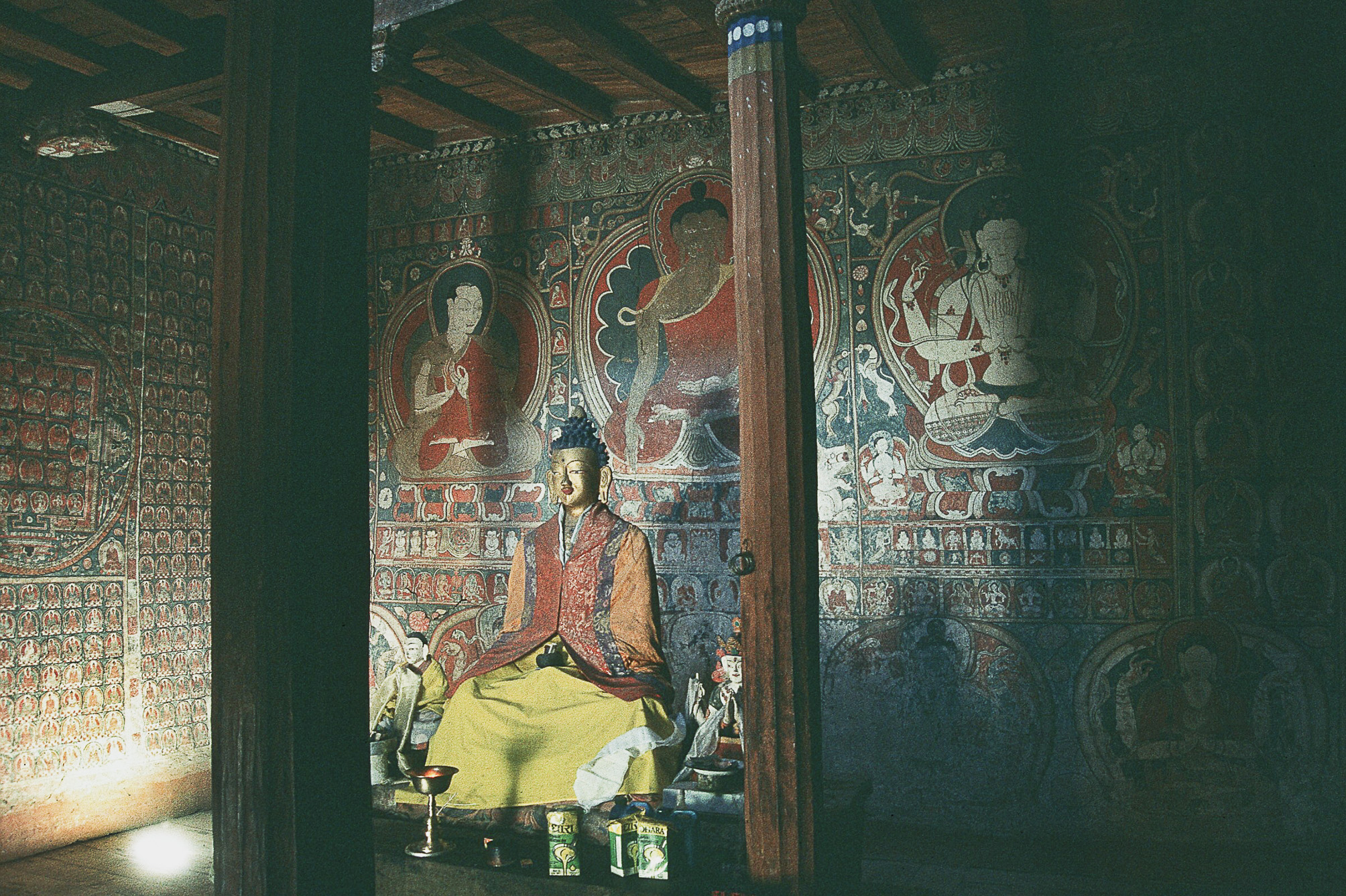 Seated Buddha statue and mural depicting three main deities illuminated from below in dark, columned hall