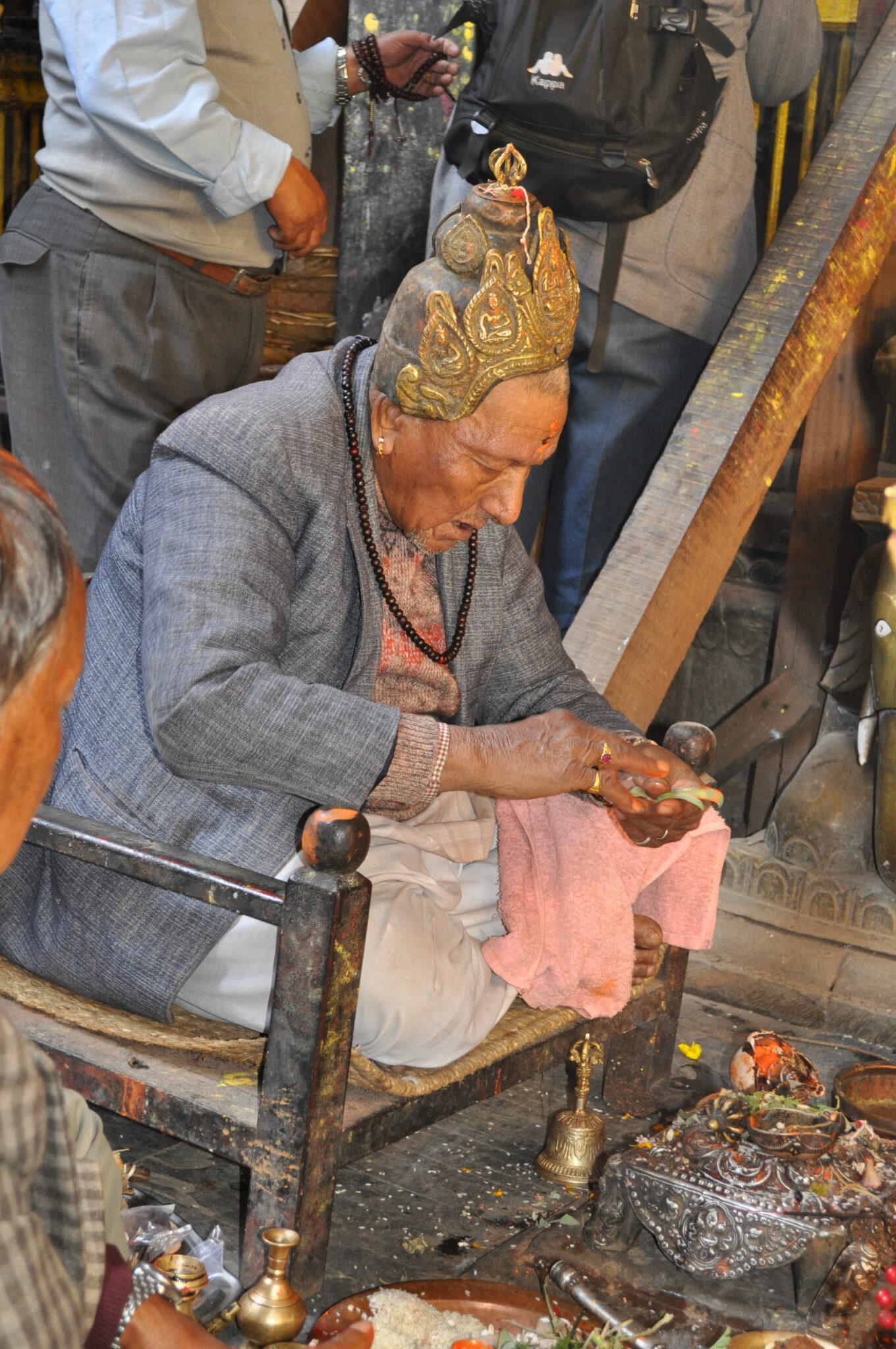 Priest wearing golden crown sits cross-legged on armchair and inclines upper body toward sacred implements on floor