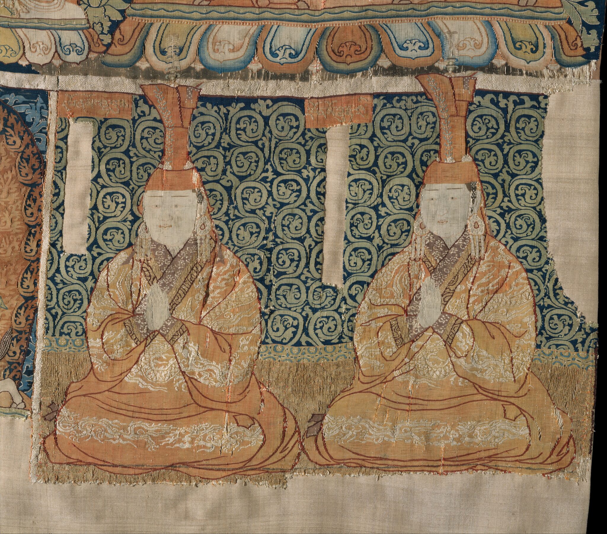 Two women dressed in saffron robes and headdresses kneeling in prayer before scrollwork background