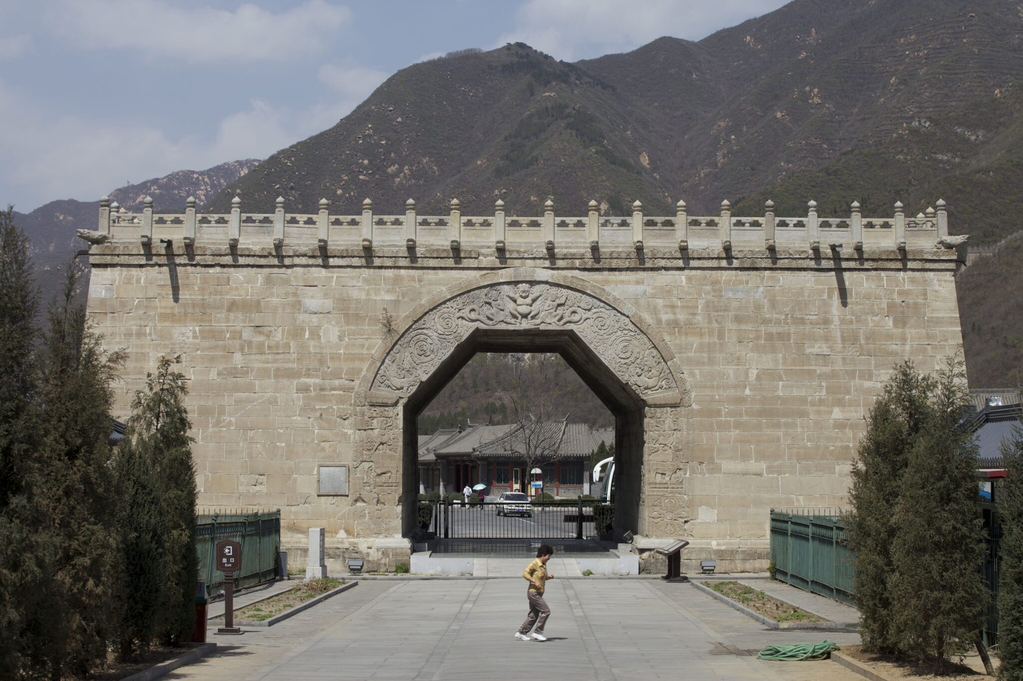 Rectangular beige stone building featuring decorative roofline and monumental arch straddling roadway; situated before forested mountainside