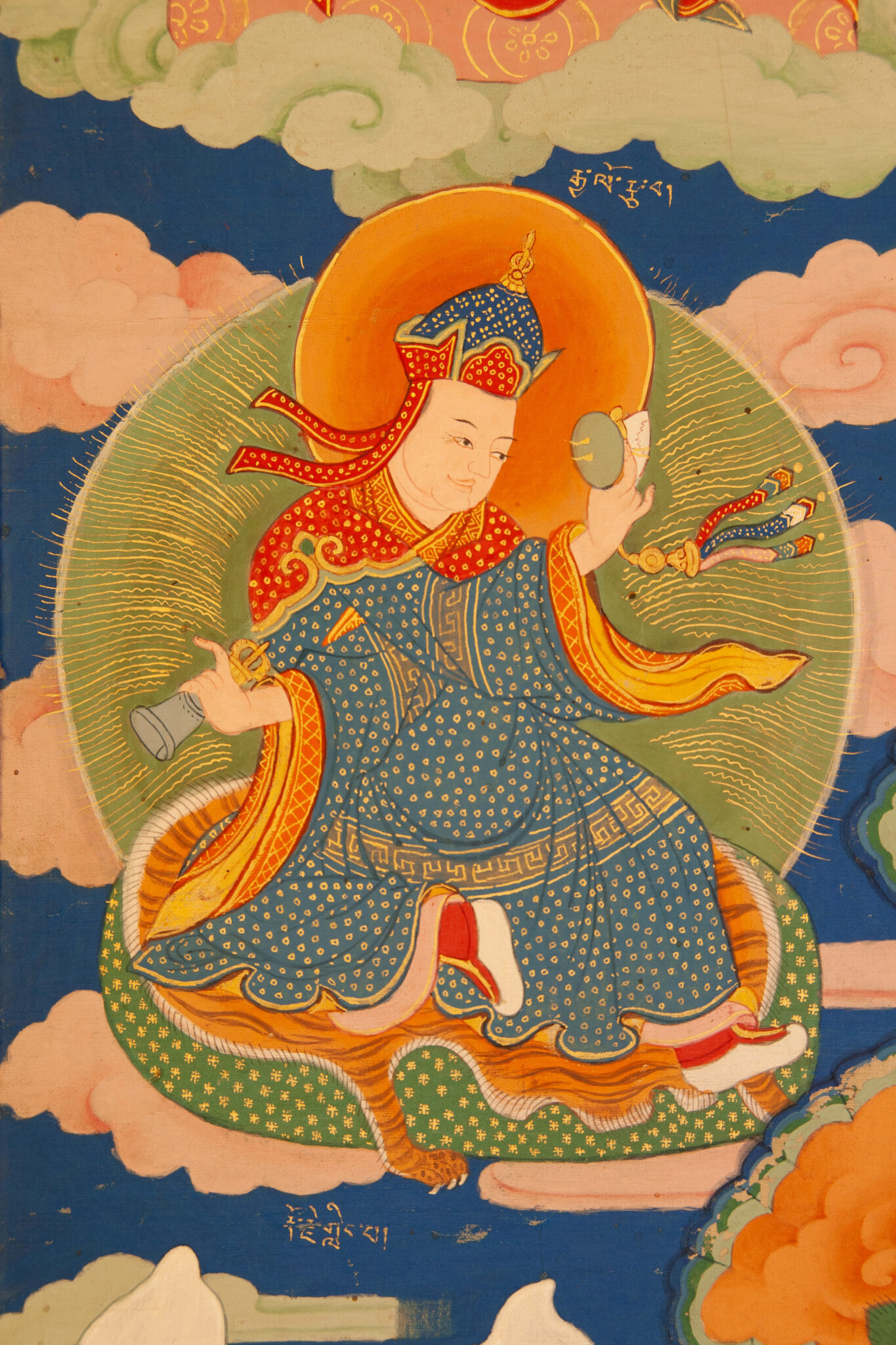 Figure dressed in gold-dotted blue robe holds religious implements in crossed arms while seated on tiger pelt before cloudy background