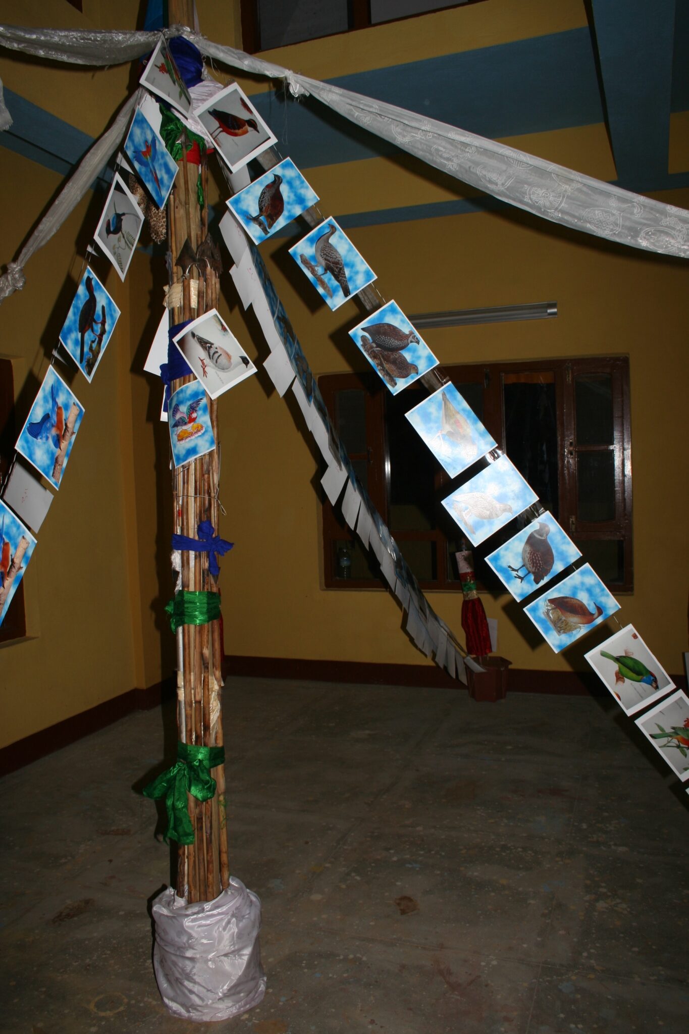 White prayer scarves and postcards featuring images of birds hang from peak of spire in center of yellow room