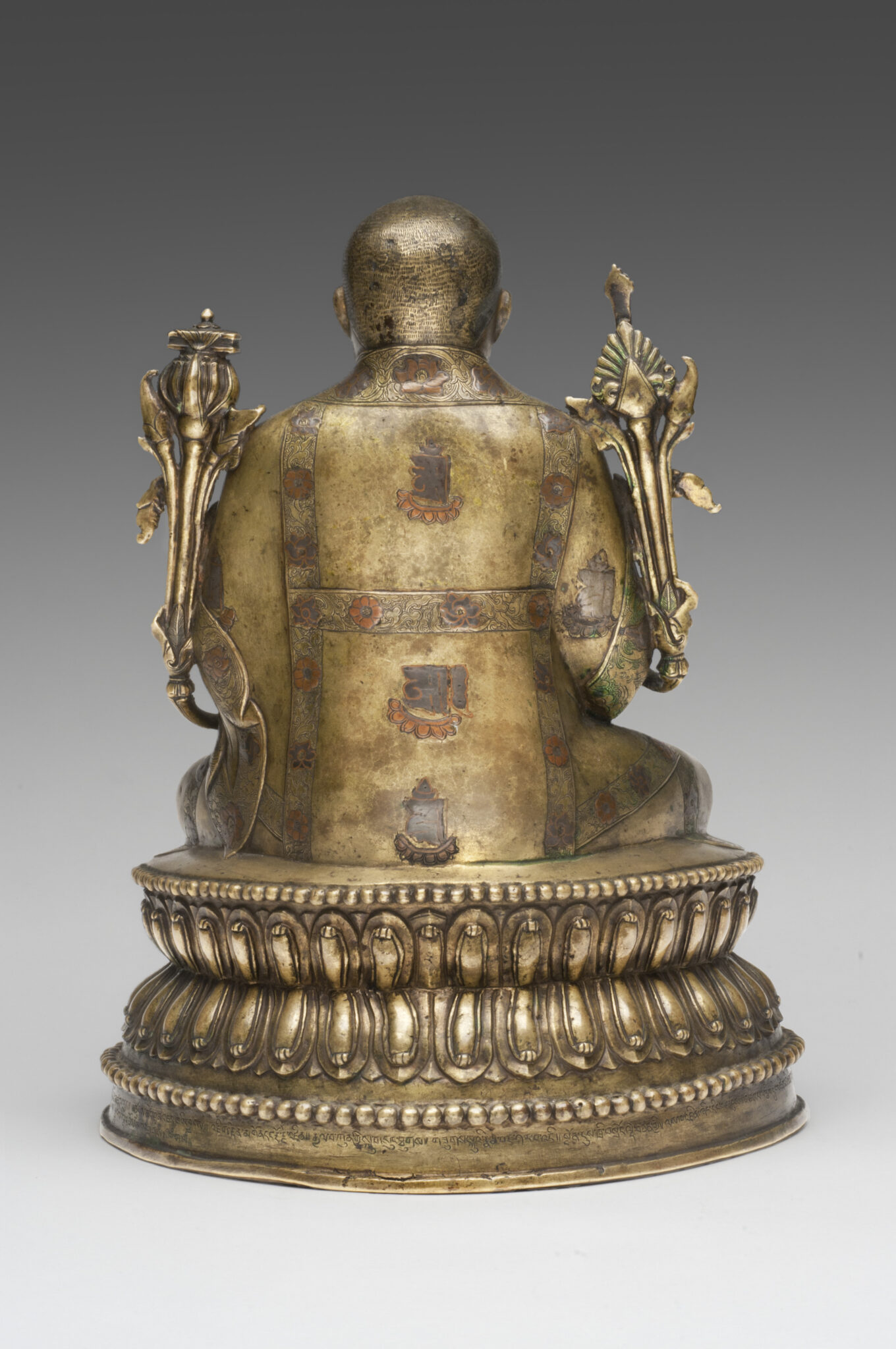 Back view of bronze-colored sculpture depicting seated figure flanked by long-stemmed blossoms