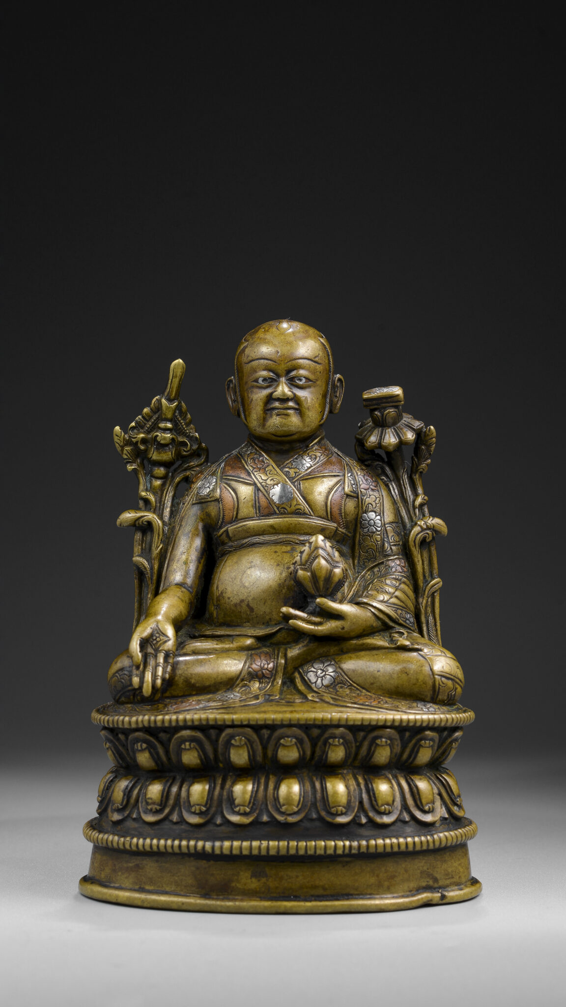 Bronze statuette depicting seated Teacher flanked by long-stemmed blossoms holding implement in right hand