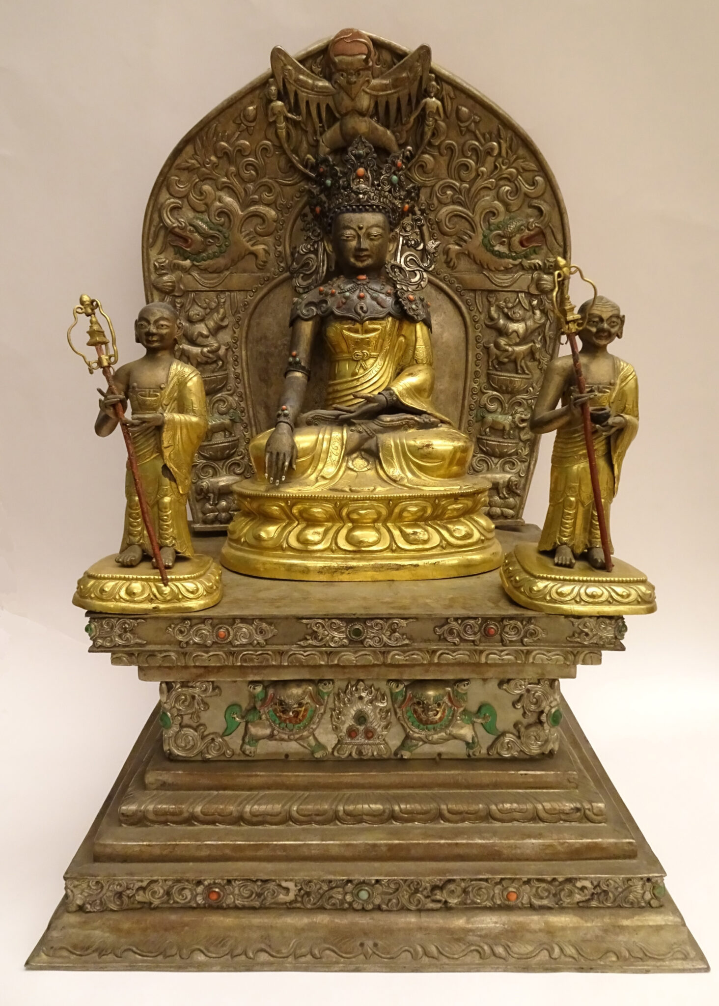 Gold- and bronze-colored sculpture depicting seated, crowned Buddha flanked by two attendants