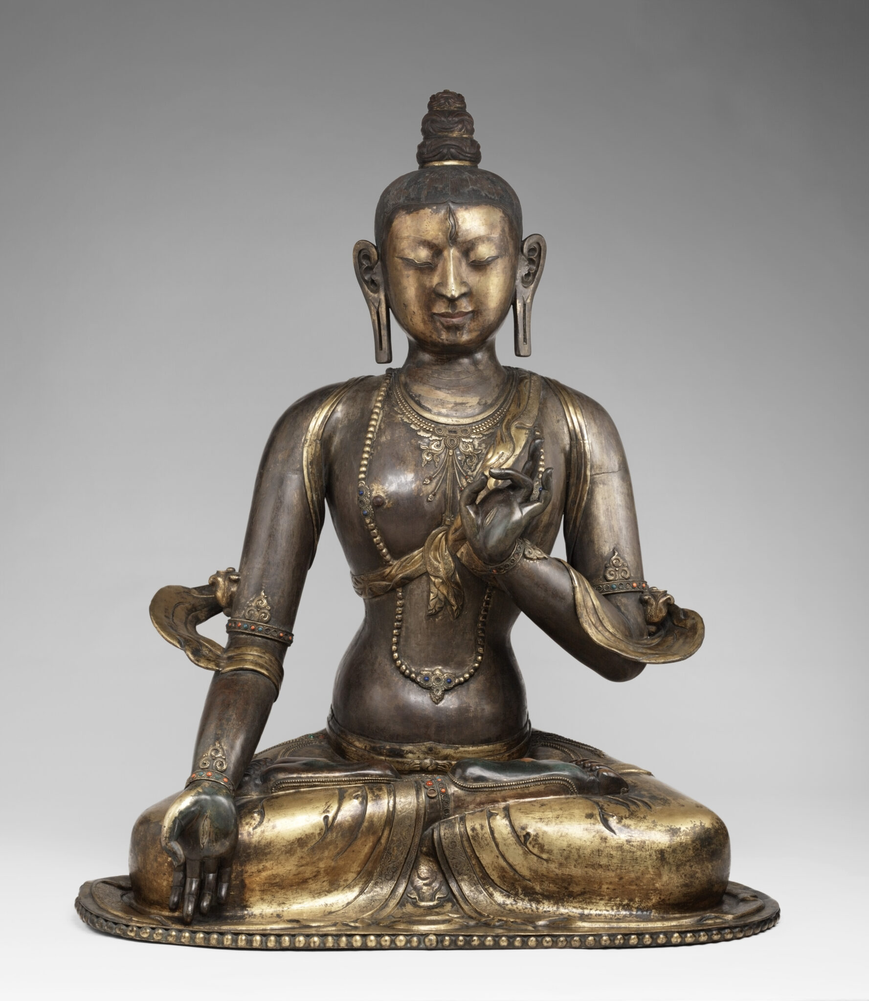 Bronze-colored statue depicting seated Bodhisattva with elongated earlobes and fluttering sash at elbows