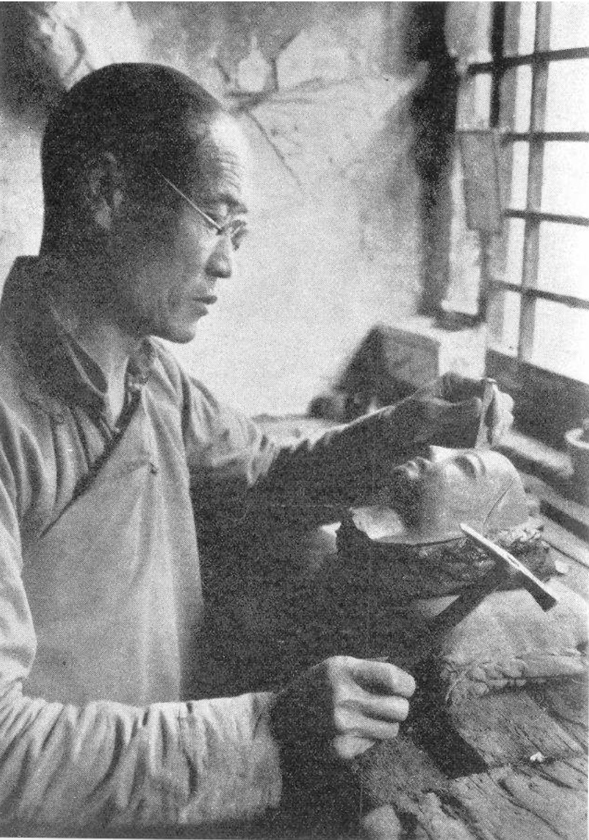 Black and white photograph of craftsman in his studio chiseling small piece of material to create face