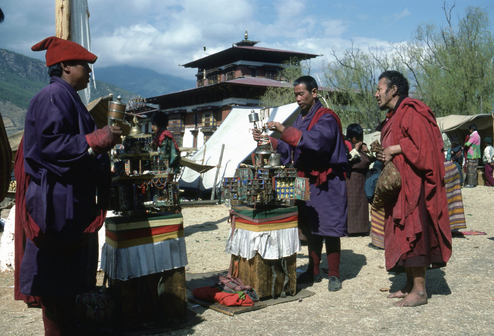 Three men in purple and red robes gather around religious implements situated on pedestals outside of pagoda-roofed building