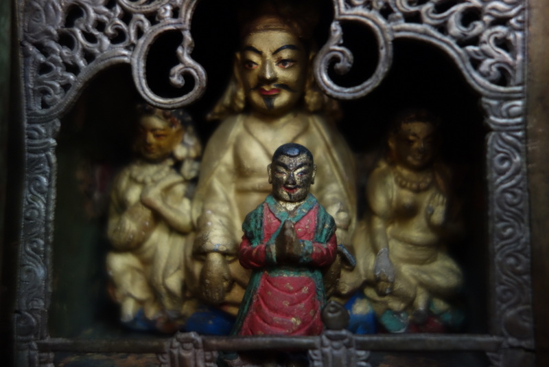 Figure wearing red and green robe kneels in prayer before three figures situated behind open-work proscenium screen