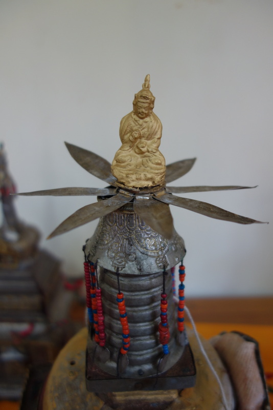 Silver-colored finial decorated with red and blue bead strings, topped by golden figure seated on blossom petals