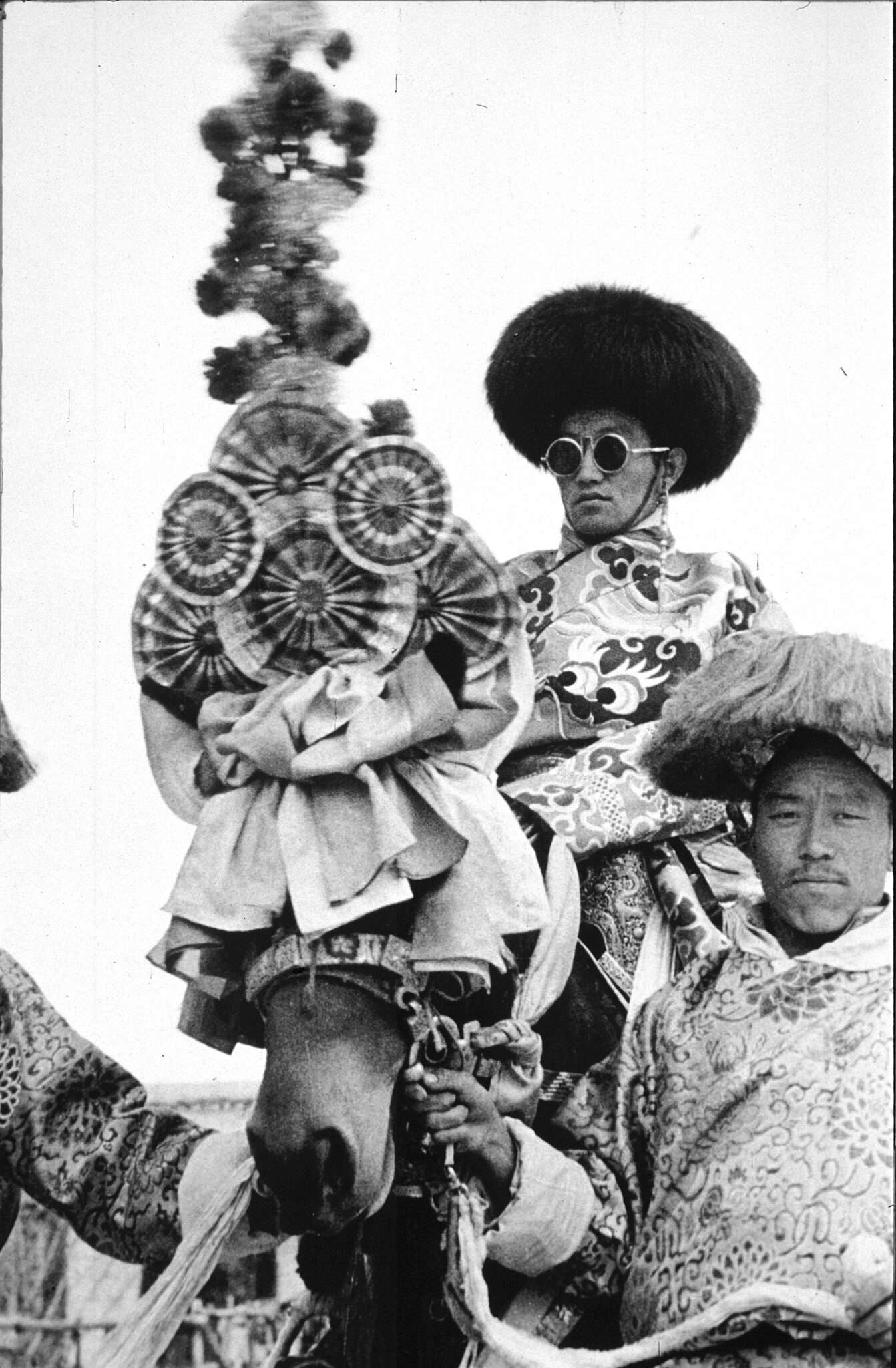 Black and white photograph of man in wide-brimmed fur hat riding horse with ornamented headdress