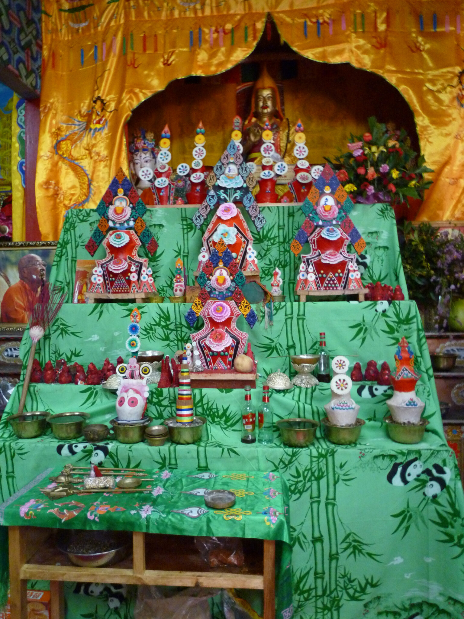 Wide view of tiered arrangement of butter sculptures and votive offerings; deity statue under golden curtain in background