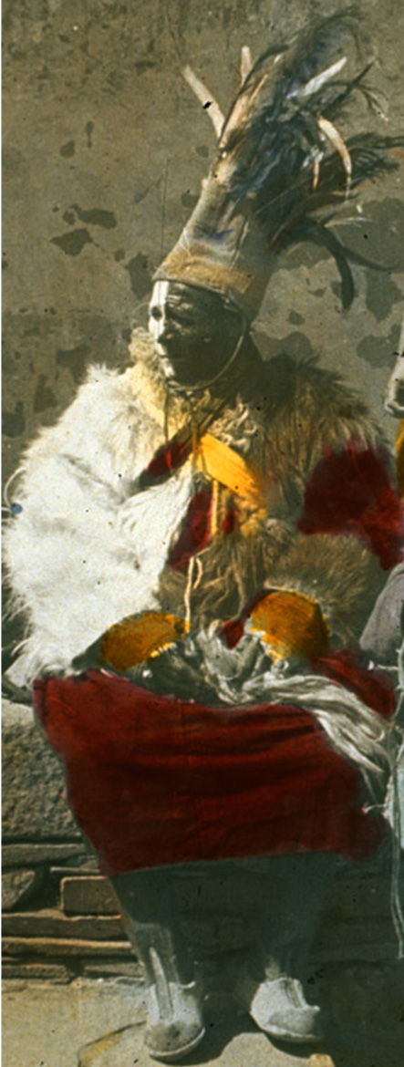 Colorized lantern slide depicting seated man dressed in red tunic, white shawl, and feathered headdress