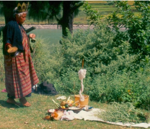 Person dressed in patterned textile and crown stands with implements before collection of devotional items