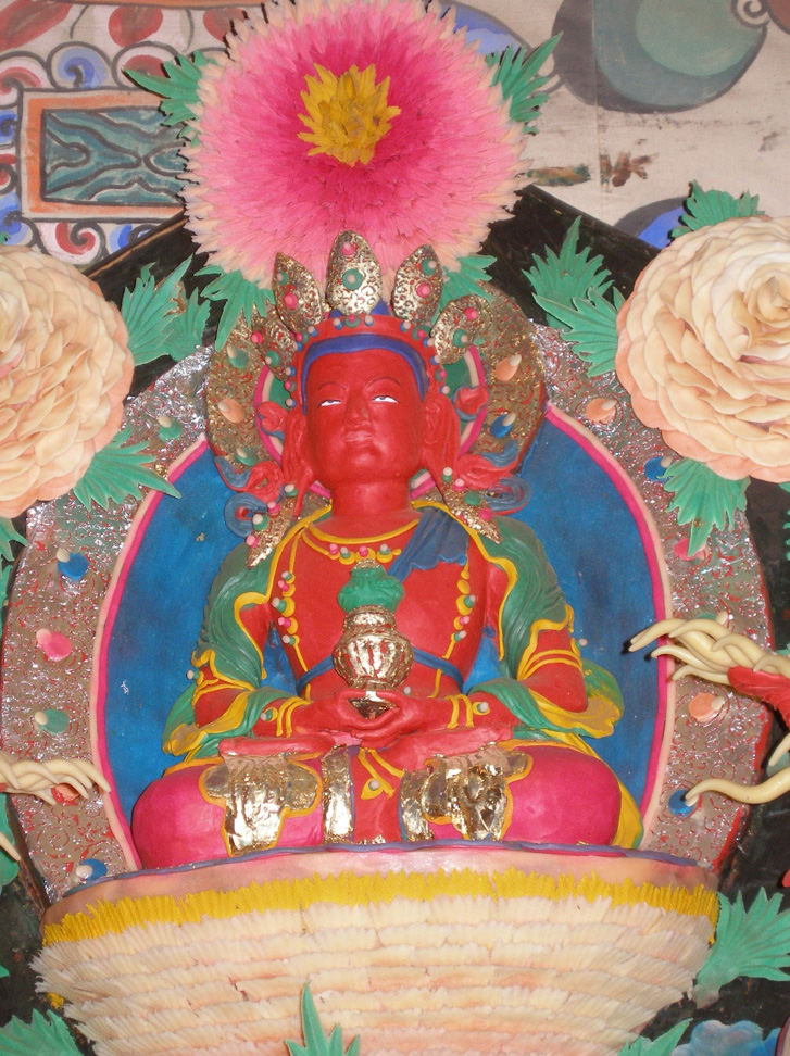 Butter sculpture depicting crowned Buddha with red skin, holding vase in hands folded in meditation