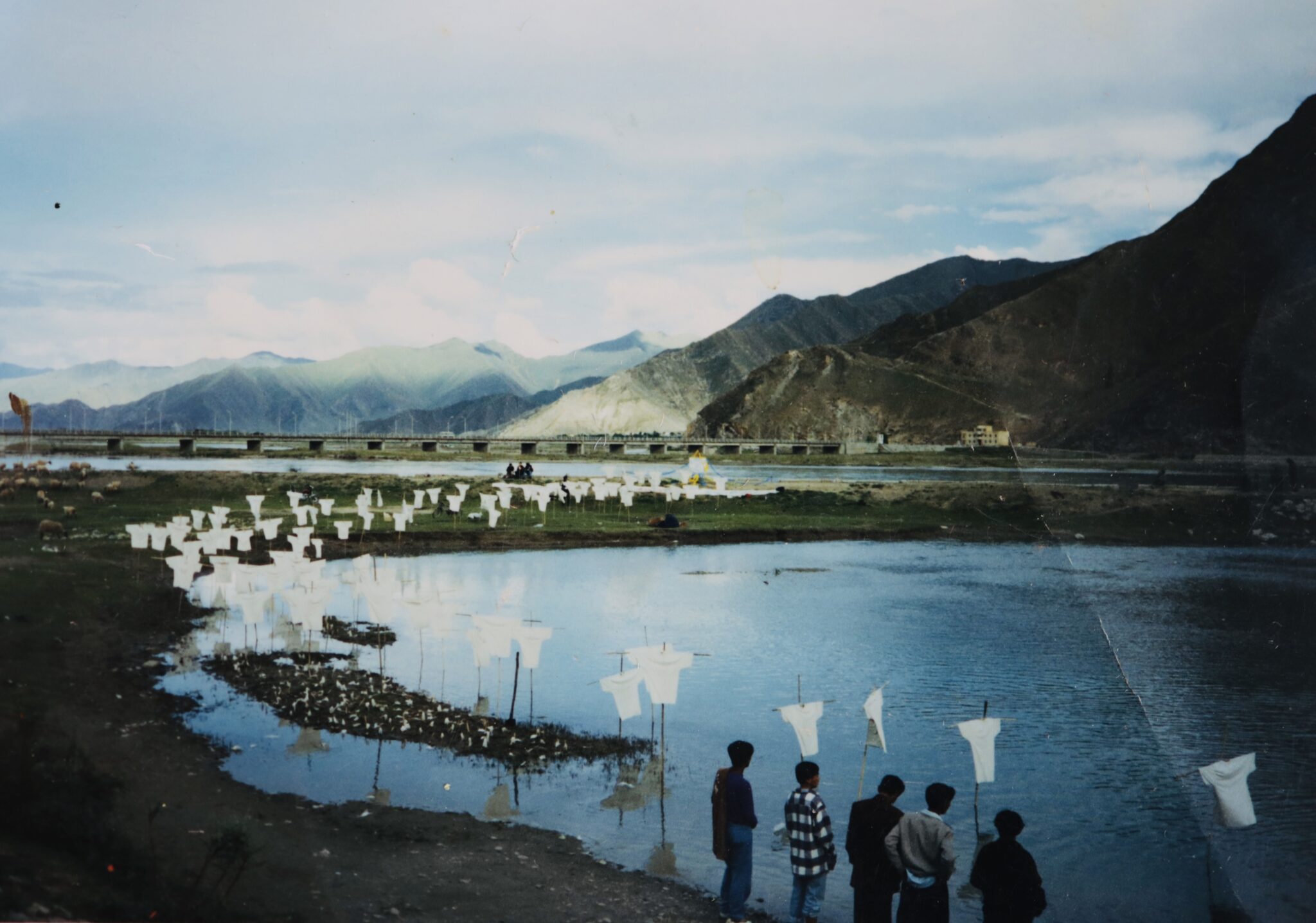 Photograph of t-shirt-shaped objects mounted on poles at shore of body of water before mountain range