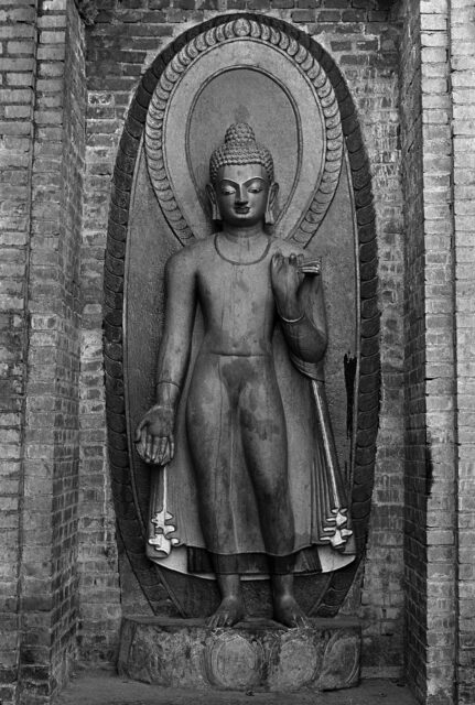 Black and white photograph of Buddha standing with right hand raised in mudra situated in brickwork niche