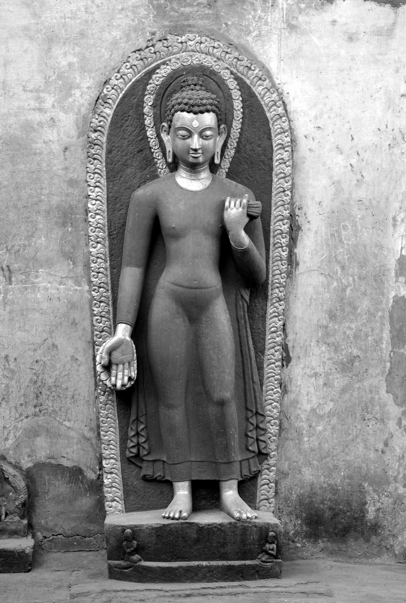 Black and white photograph of sculpture depicting haloed Buddha wearing sheer tunic standing before full-body nimbus