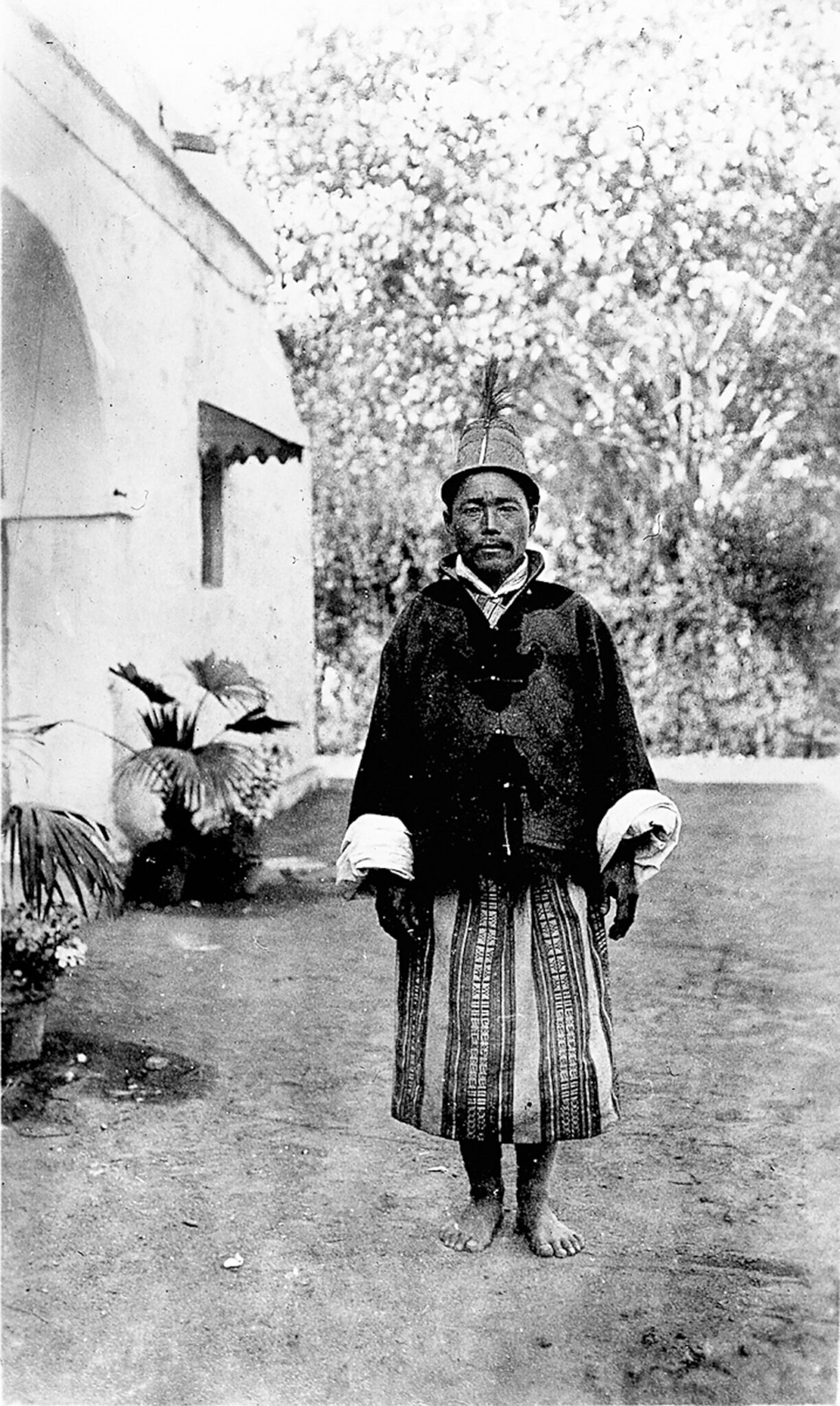 Black and white photograph of man wearing dark jacket and patterned dhoti standing beside building