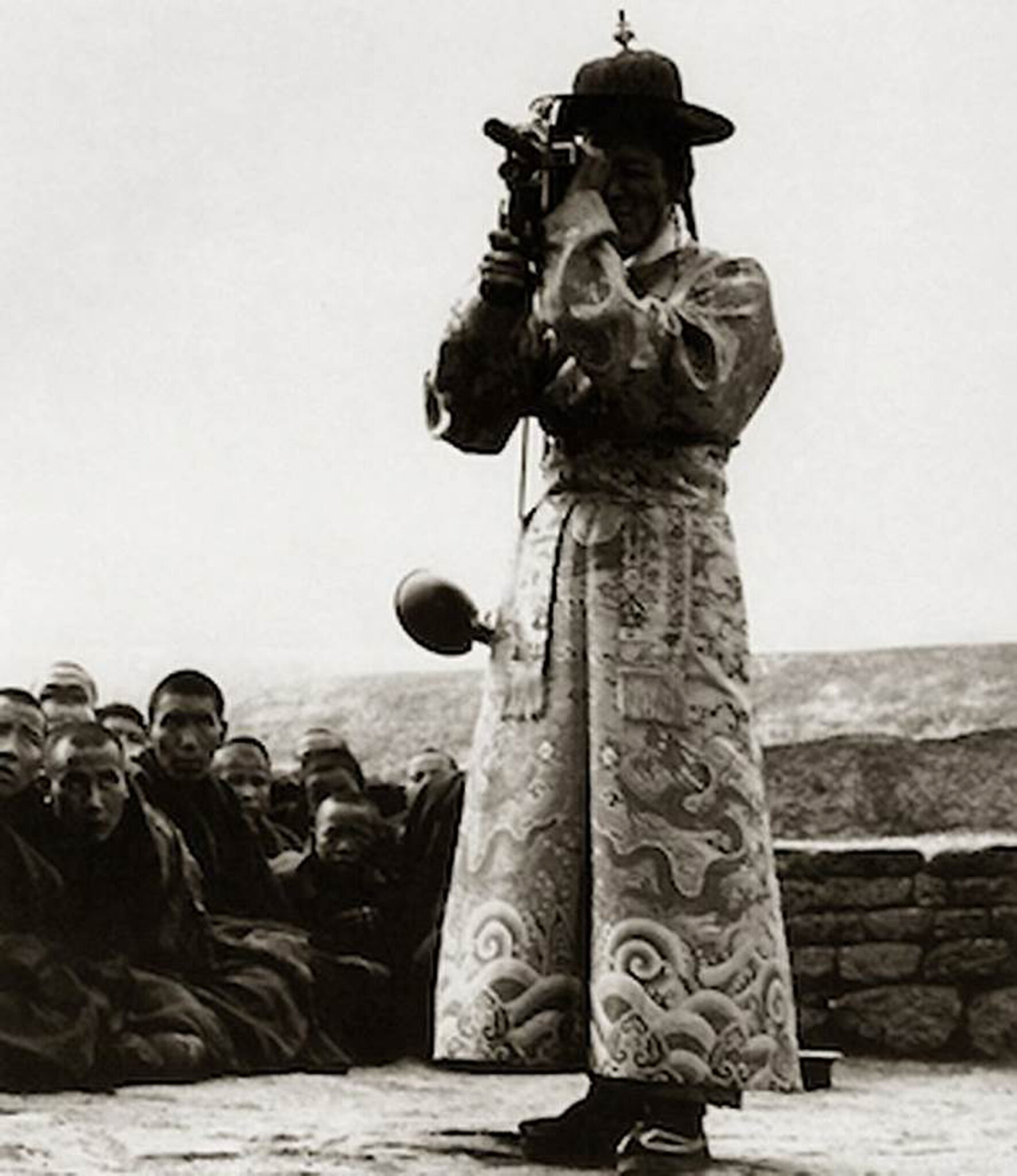 Black and white photograph of man wearing embroidered robe holding camera up to eyes beside monks seated at left