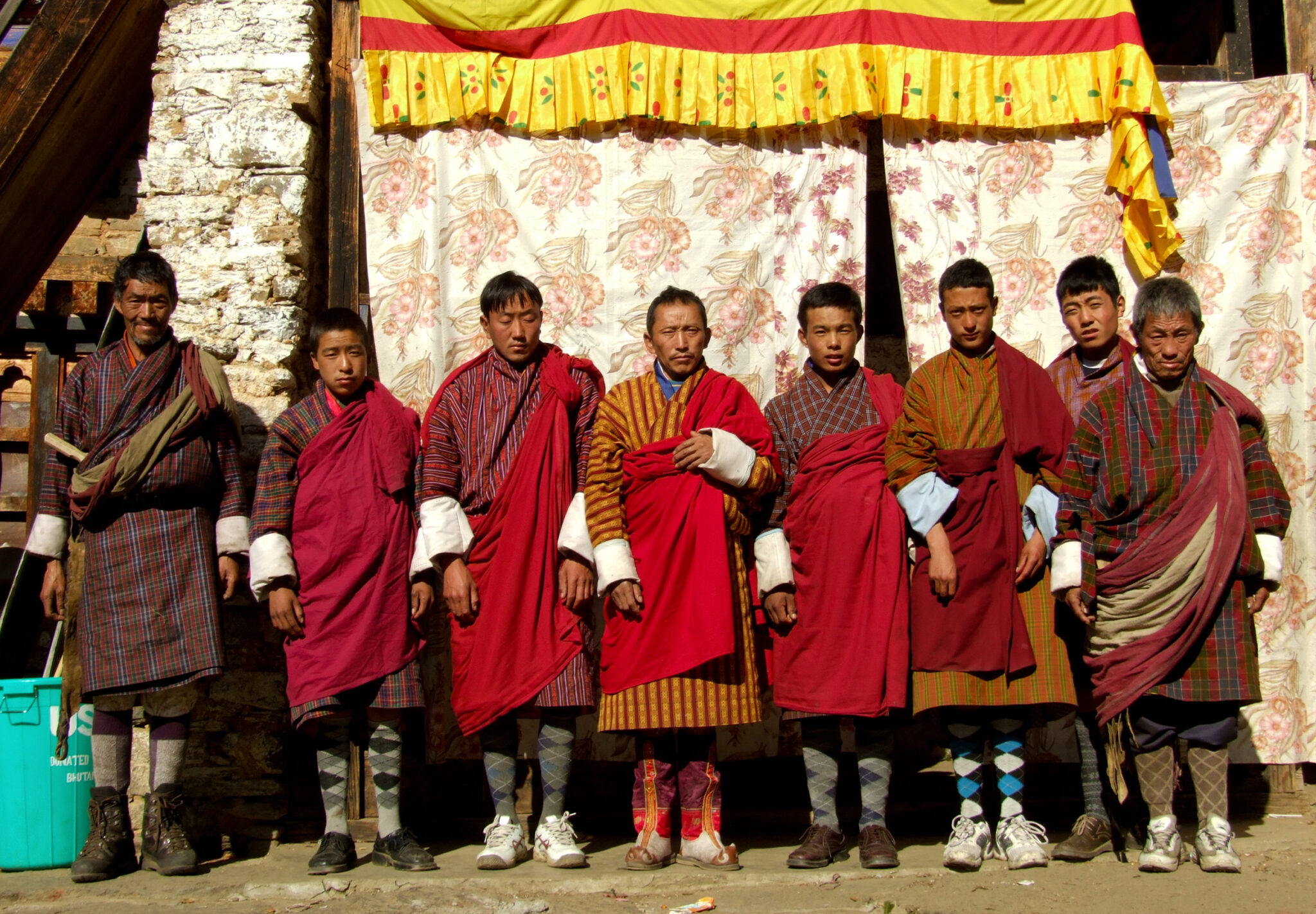 Eight men stand in line wearing red garments draped over plaid tunics, which come just below the knees