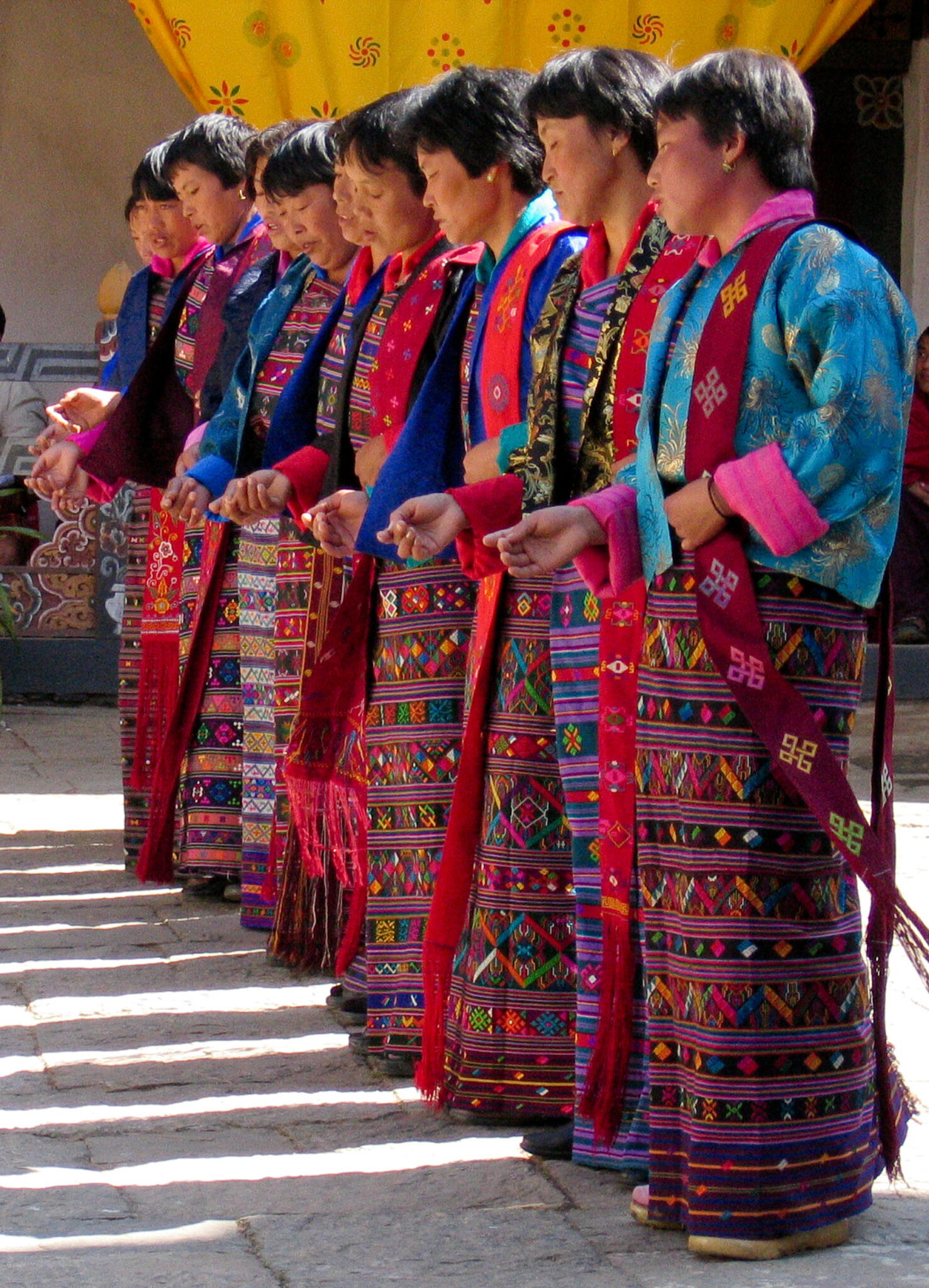 Women stand in line wearing blue embroidered jackets and floor-length skirts woven in multitude of colors