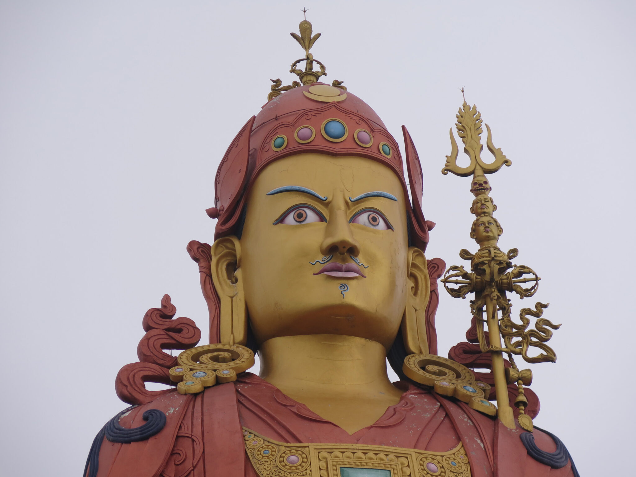 Head and chest of colossal statue depicting stern-faced Guru with vajra staff at right shoulder