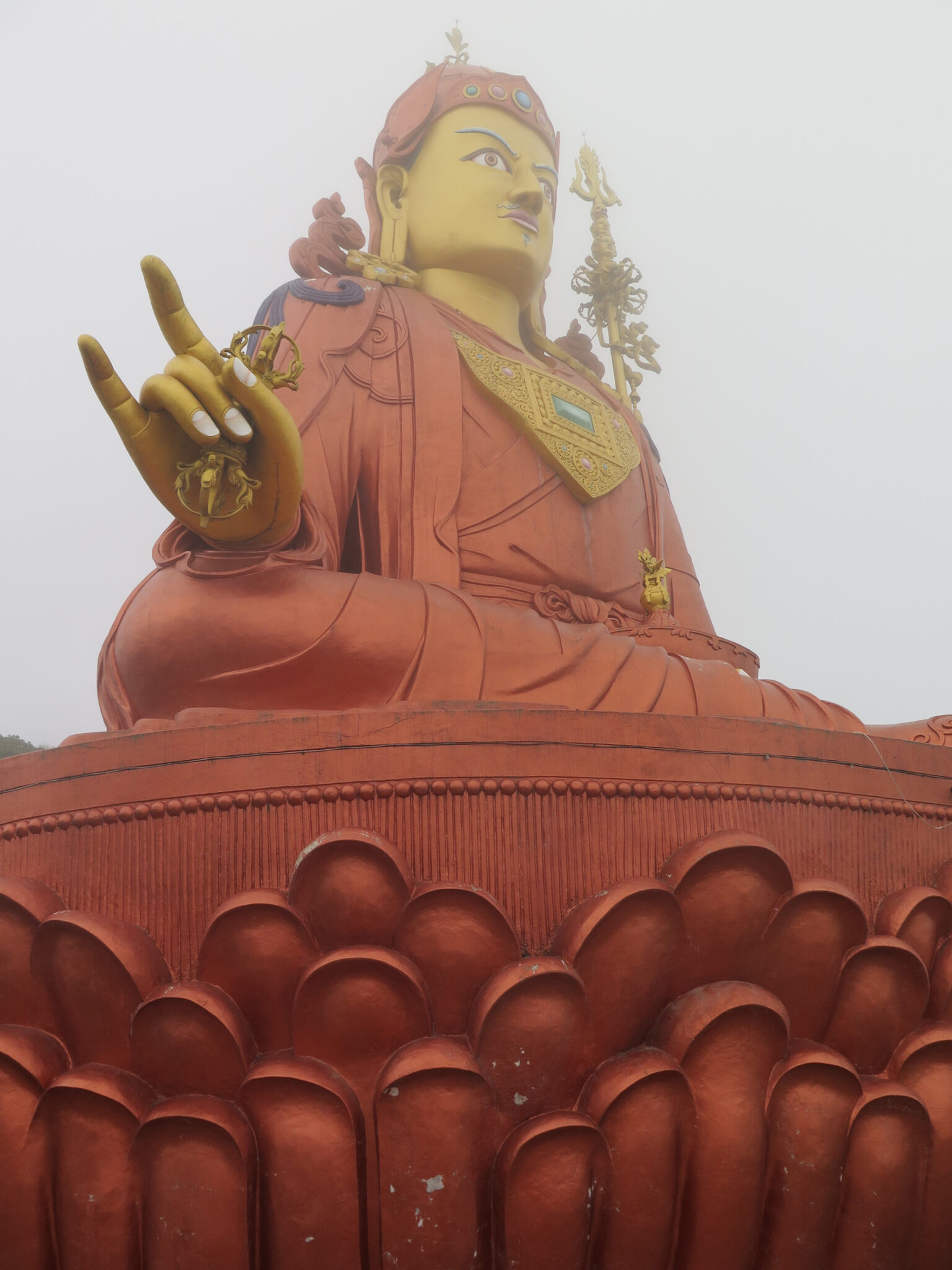 View from below of mist-shrouded colossal statue depicting Guru seated on lotus pedestal