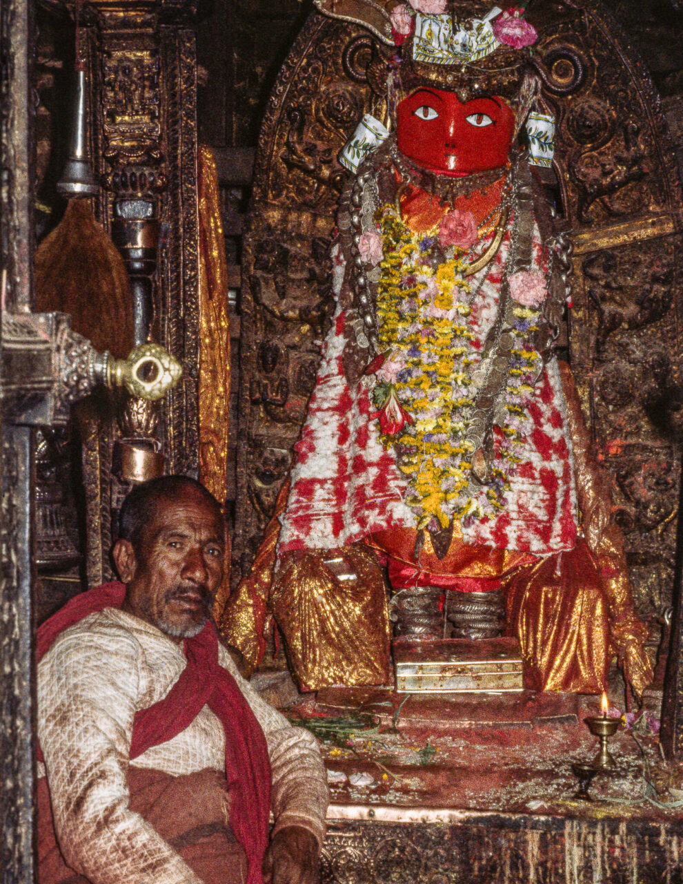 Man sits before statue adorned with garlands and textiles; statue has red, abstract face