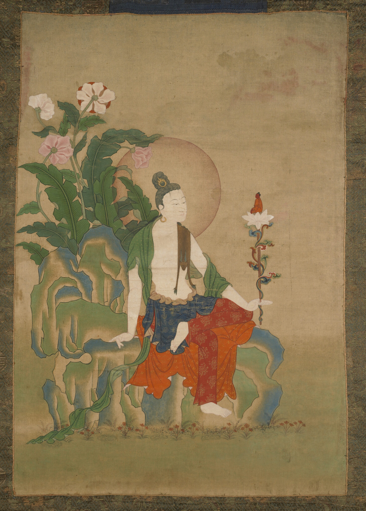 Bodhisattva dressed in sumptuous dhoti holds blossom in right hand while seated on rocky outcropping