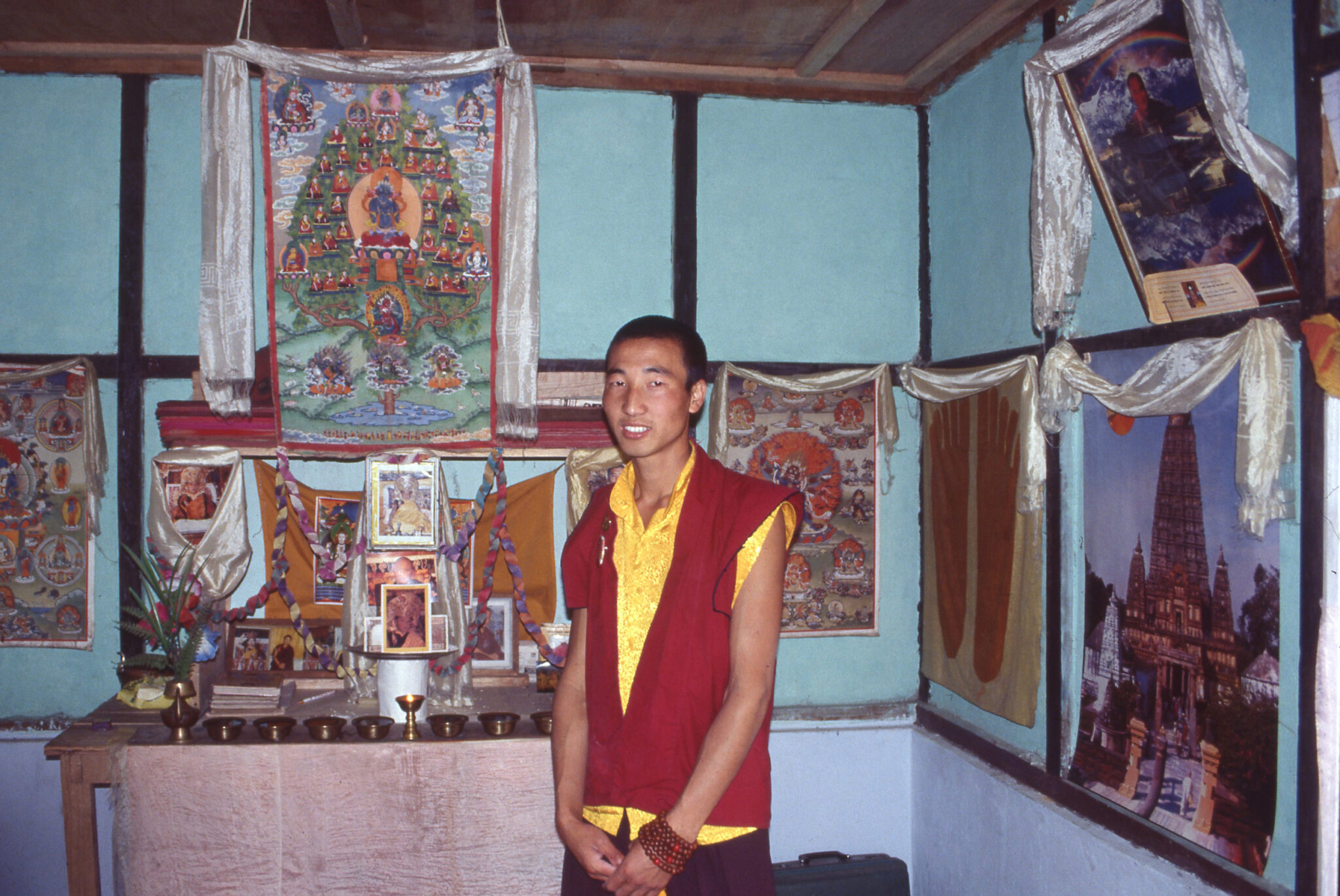 Man stands in corner of room; walls hung with religious artworks and photographs adorned with white scarves