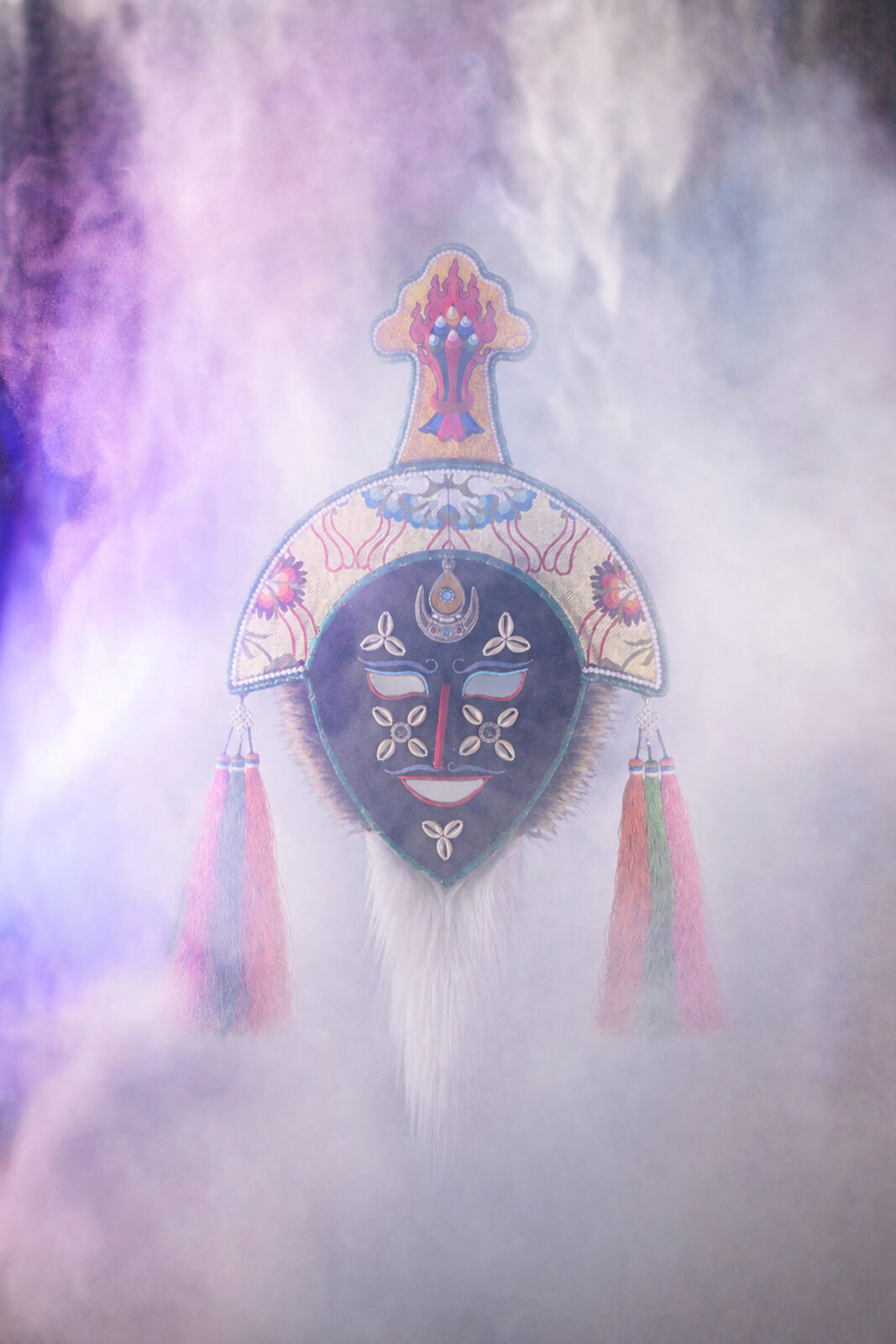 A colorful mask floating in the air behind light purple and blue smoke