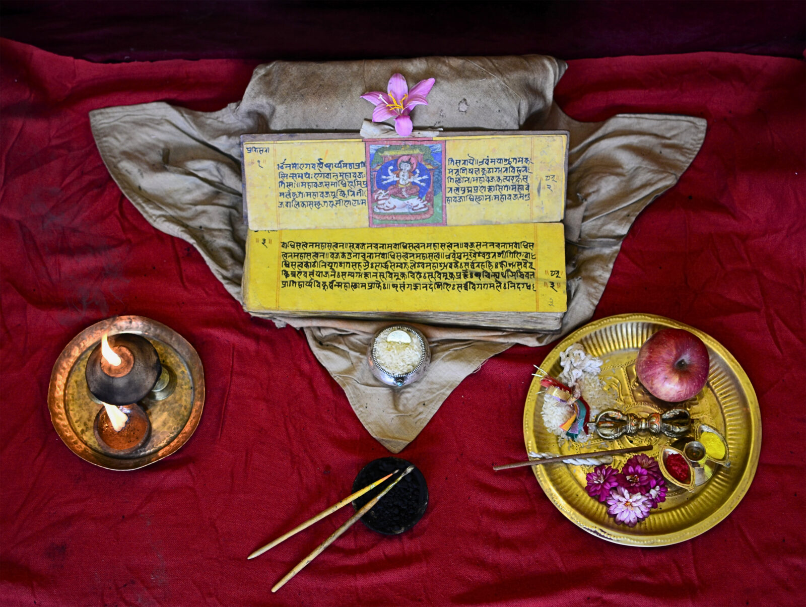 Two yellow strips of paper with writing on a beige cloth over a red cloth, with a dish of two small candles to the left, a dish of two paintbrushes below, and a dish of an apple and flowers to the right
