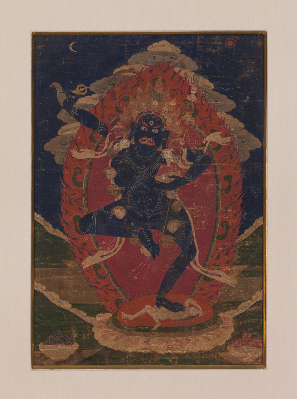 Thangka painting of a black wrathful multi-armed deity dancing within an orange flame aureole, standing on a prone figure, against a dark blue night sky with a crescent moon and sun.
