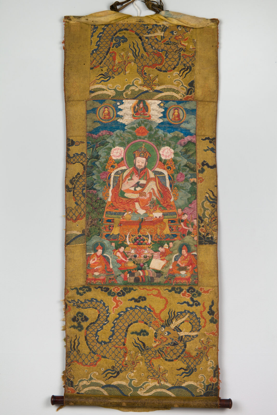 Painting of large seated figure with colorful robes surrounded by smaller figures above and below mounted on vertical strip of golden patterned fabric.