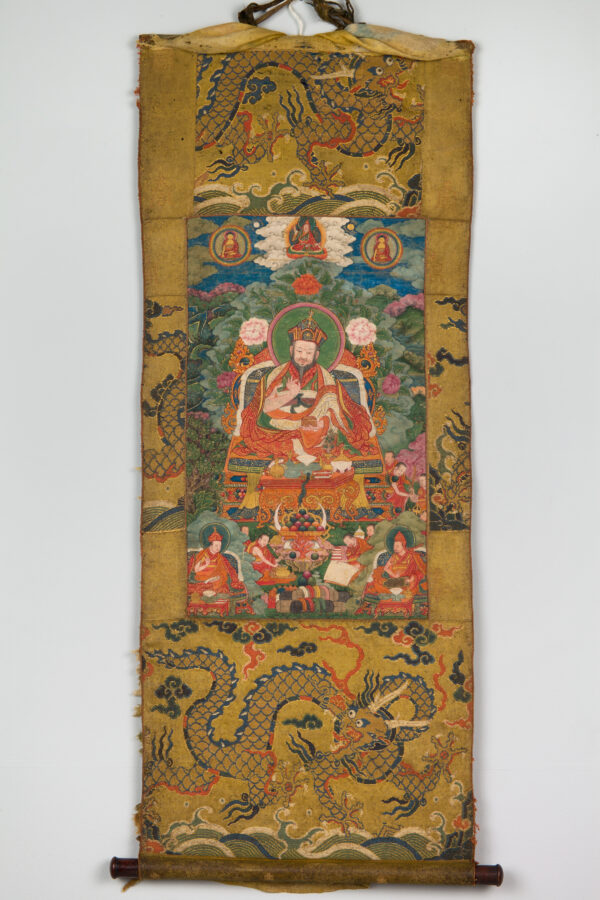 Painting of large seated figure with colorful robes surrounded by smaller figures above and below mounted on vertical strip of golden patterned fabric.