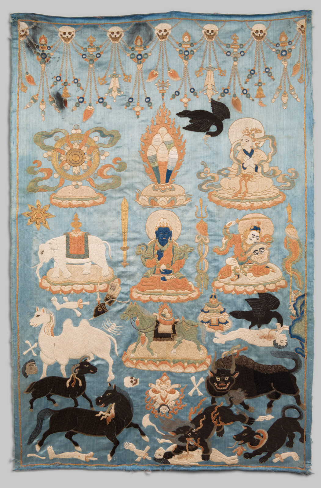 Embroidered white and black animals, two black ravens, and human figures populate a light blue background. Skulls and beaded strings decorate the upper edge, with black animals in the lower portion. A dark, blue-skinned figure sits at center, one of three human figures. There are seven animals, including a white Bactrian camel and a black snow lion devouring a corpse. All represent symbolic offerings.