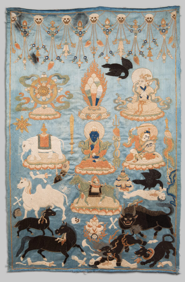 Embroidered white and black animals, two black ravens, and human figures populate a light blue background. Skulls and beaded strings decorate the upper edge, with black animals in the lower portion. A dark, blue-skinned figure sits at center, one of three human figures. There are seven animals, including a white Bactrian camel and a black snow lion devouring a corpse. All represent symbolic offerings.