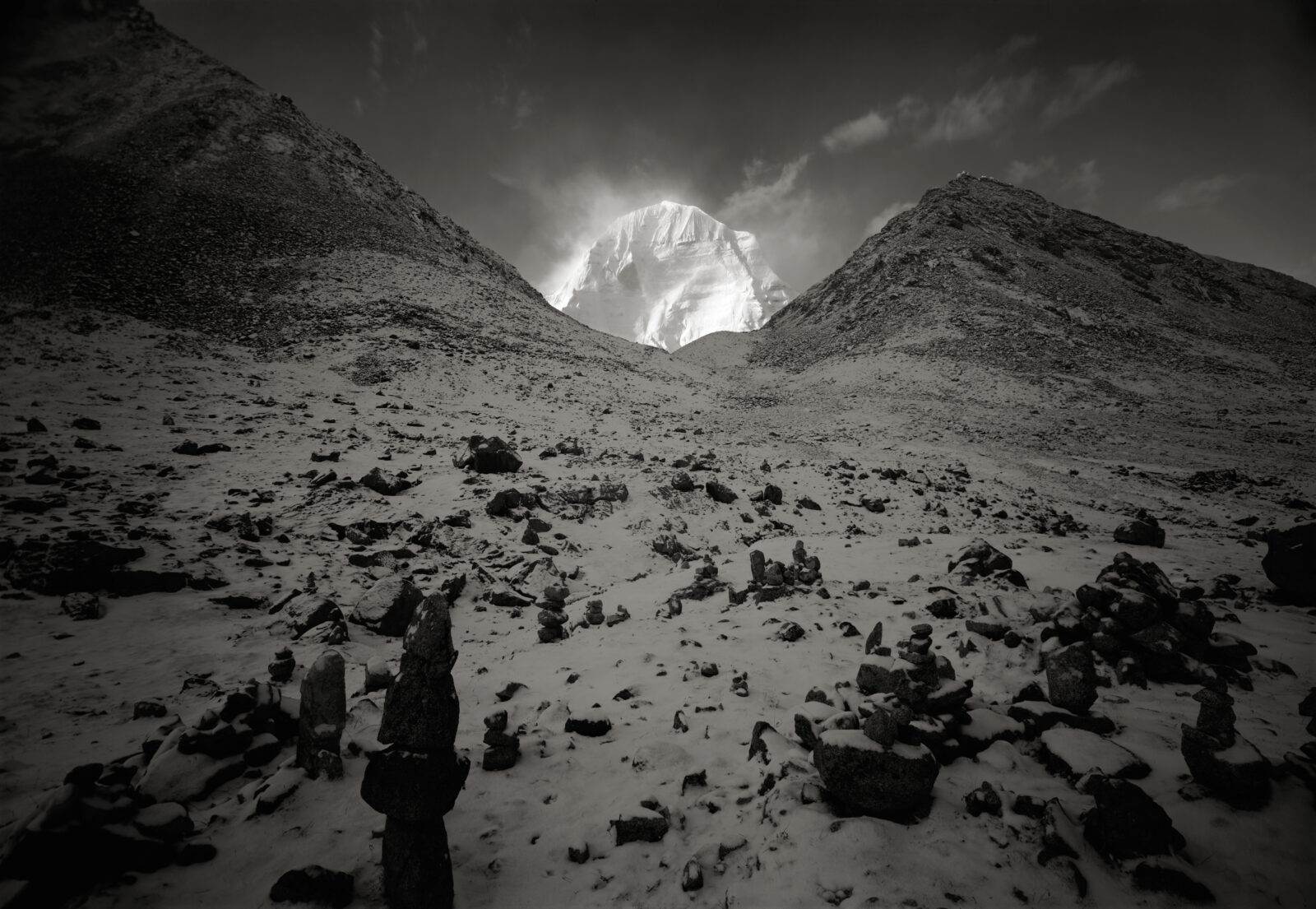 Black-and-white image of bright white snow-covered mountain rising from valley between two darker peaks lightly covered with snow.