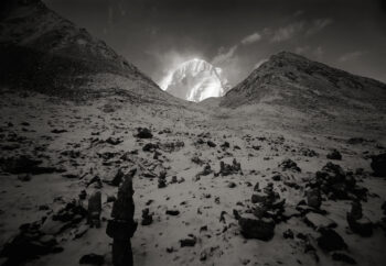 Black-and-white image of bright white snow-covered mountain rising from valley between two darker peaks lightly covered with snow.