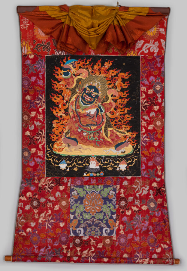 Thangka scroll painting of a wrathful flame-wreathed deity seated on a lotus base against a black background.