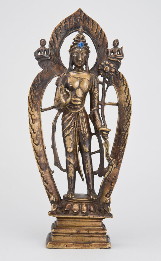 Bronze statue of a standing bodhisattva with a blue gemstone in the crown, holding a lotus stem, framed by an arched flame- patterned aureole with two small figures at the top corners, who sit on a rectangular base.